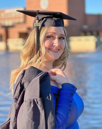 一名女子戴着毕业典礼帽，把长袍披在肩上，站在德克萨斯大学A分校的加文湖前&M University-商务.